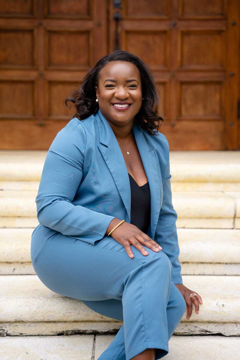 Smiling person in a blue suit sitting on stone steps in front of a wooden door.