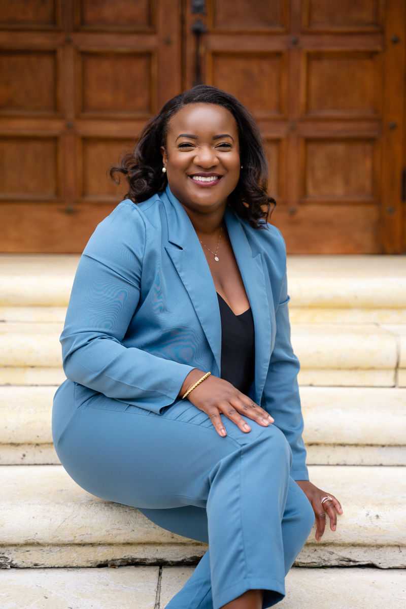 Smiling person in a blue suit sitting on stone steps in front of a wooden door.